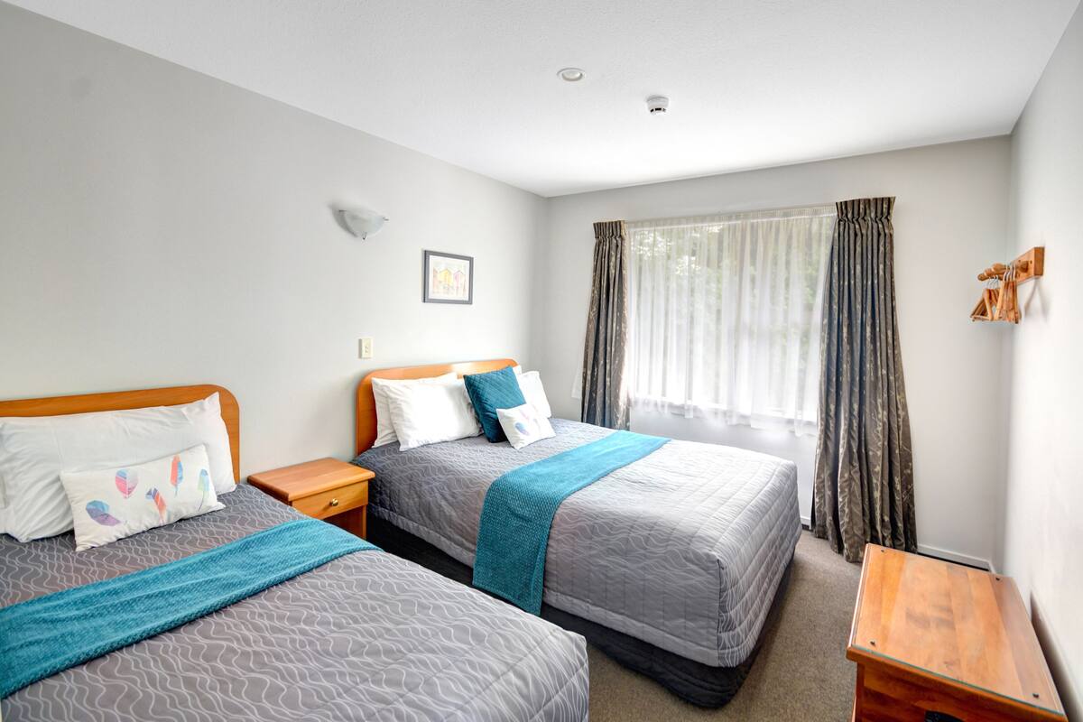 A hotel room with two beds, a wooden nightstand, a small wooden table, and a window with curtains.