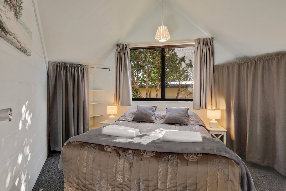 A bedroom with a bed, two bedside tables with lamps, a window with curtains, and a ceiling light.