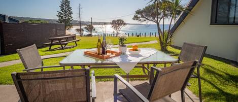 A patio with a table and chairs, overlooking a coastal view with a grassy area and a wooden fence.
