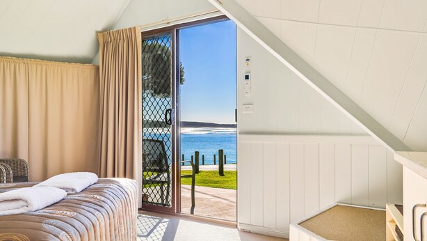 A bedroom with a bed, a window with curtains, a view of the sea, and a white wall.