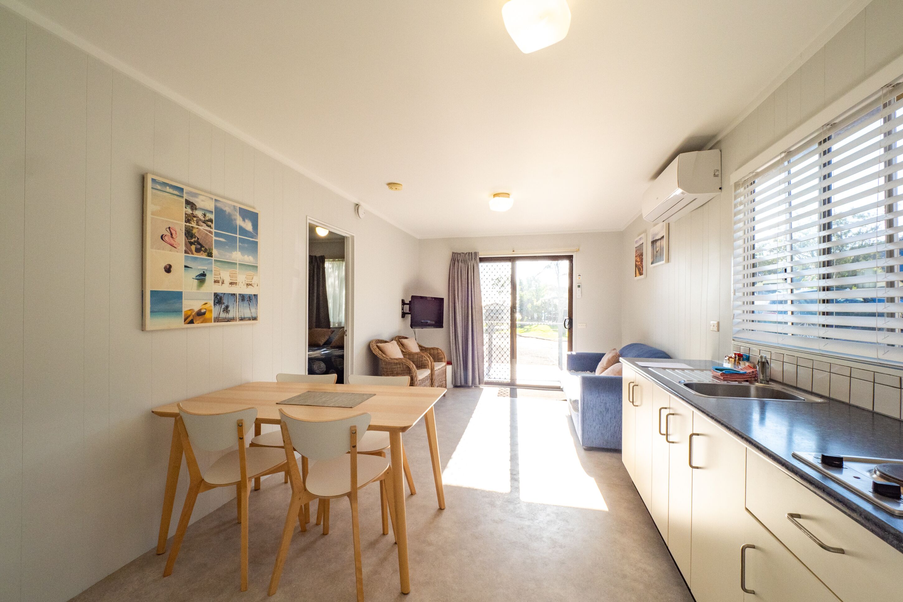 Una cocina moderna con comedor, una sala de estar con sofá y televisor, y un ventanal con persianas.