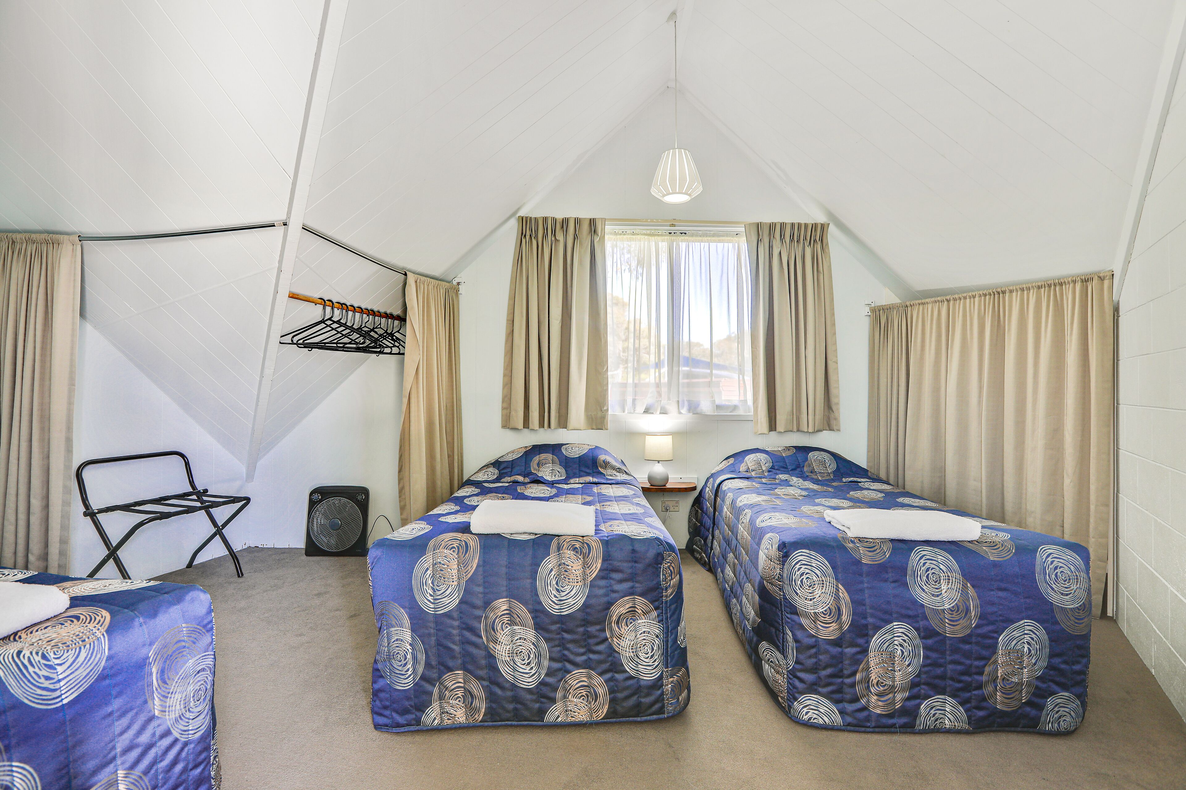 A room with two beds, a chair, a fan, and a window with curtains.