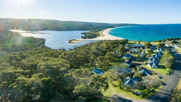 An aerial view of a coastal area with a beach, a forested area, and a row of houses with pitched roofs.