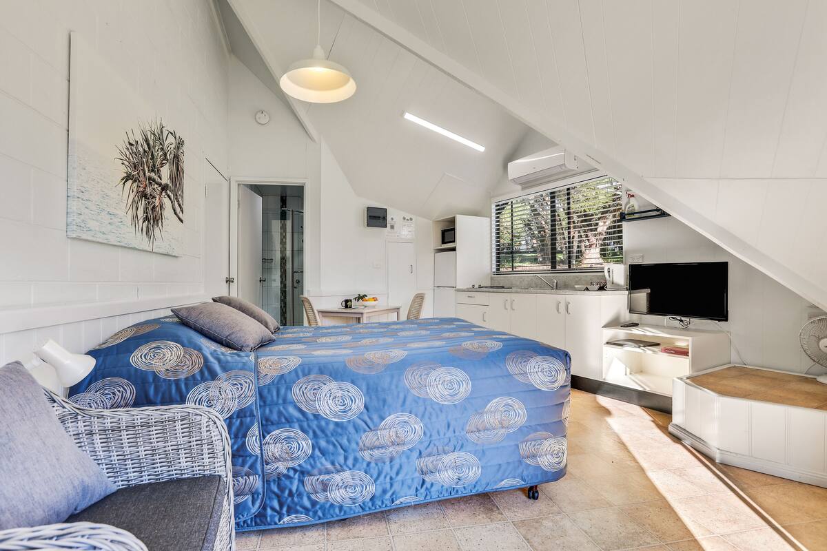 A compact bedroom with a bed, a chair, a desk, a TV, and a kitchenette.