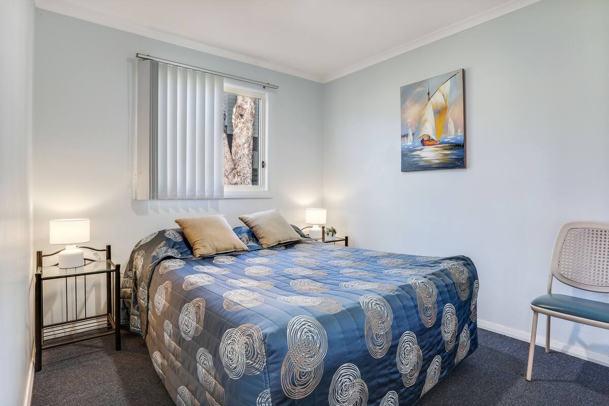 A bedroom with a bed, a chair, a window with blinds, a painting on the wall, and bedside lamps.