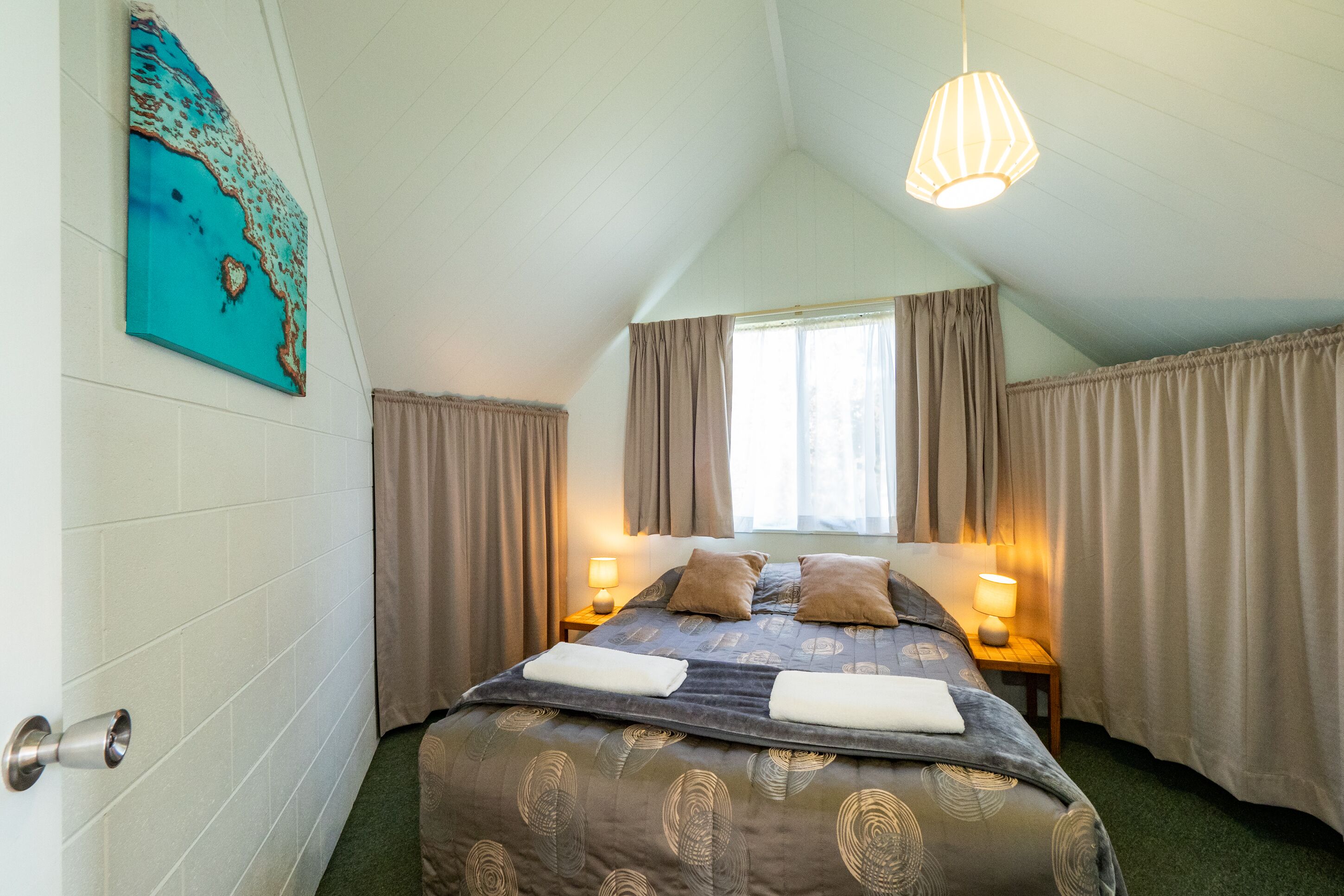 A bedroom with a bed, two bedside lamps, a window with curtains, a painting on the wall, and a door with a handle.