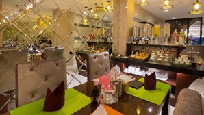 Un bufé de desayuno con diversos alimentos y bebidas, una zona de estar con sillas y mesas, y una decoración interior moderna con superficies reflectantes e iluminación colgante.