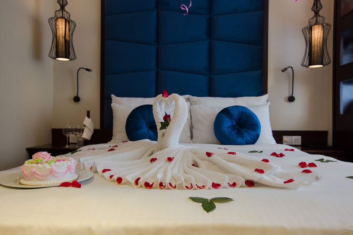 A bed with a white duvet, blue pillows, and a decorative towel arrangement with rose petals.