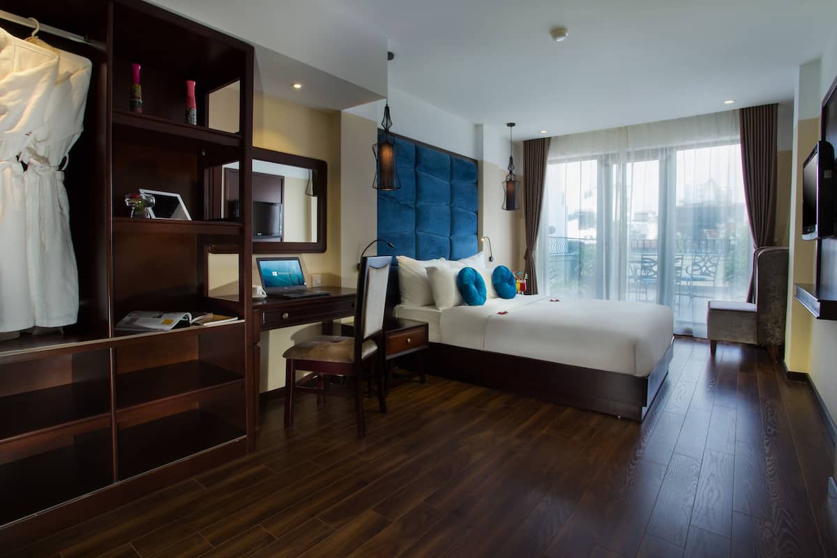 A hotel room with a large bed, a desk with a laptop, a wardrobe, and a view of the city through large windows.