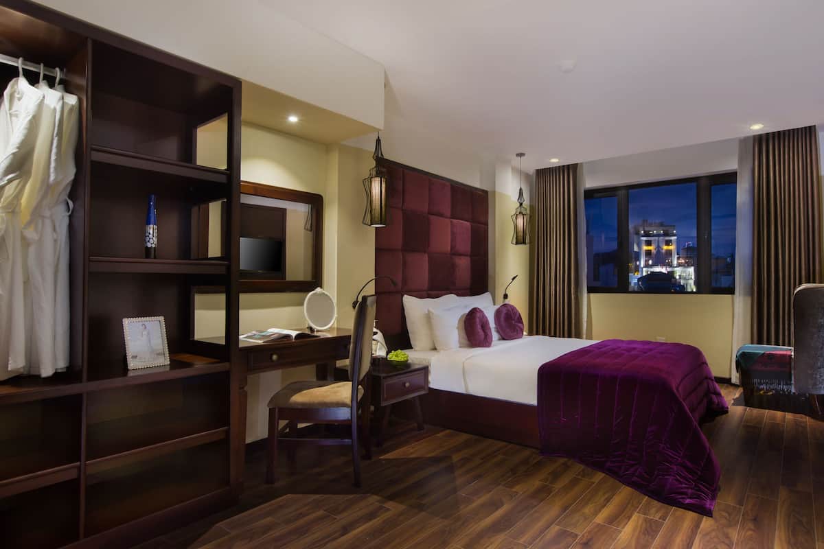 A hotel room with a large bed, a desk with a chair, a wardrobe, a mirror, and a view of the city at night.