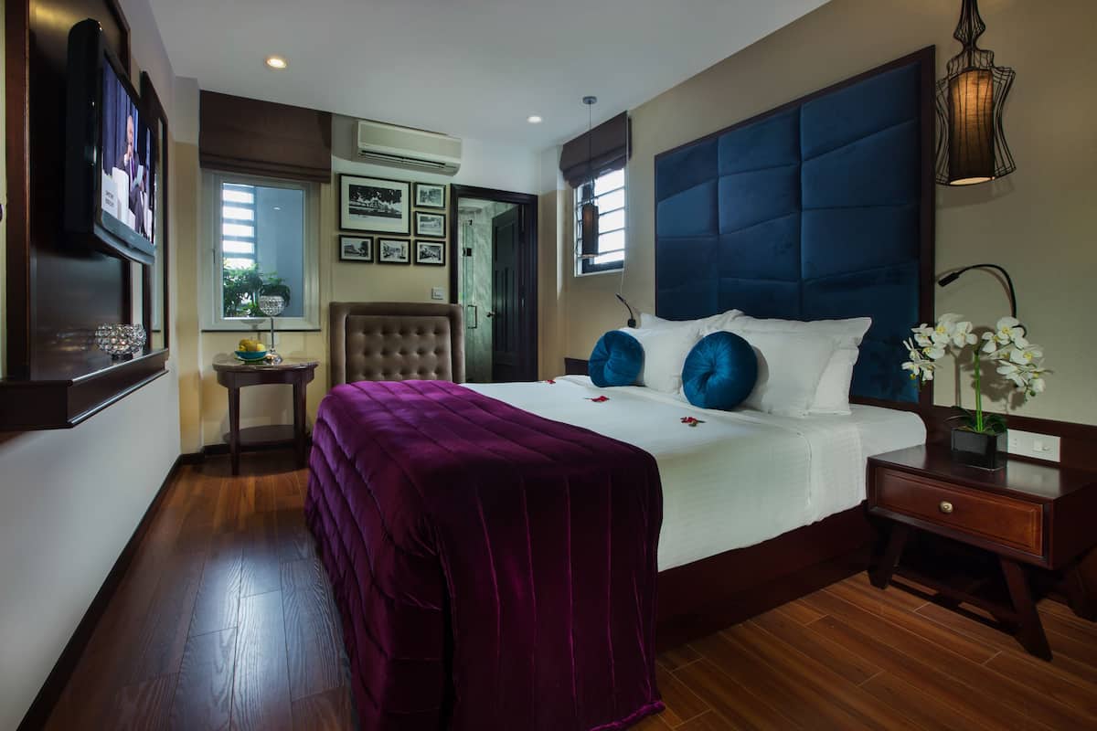 A bedroom with a large bed, a television, a chair, a nightstand, and a window with blinds.