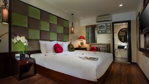 Una habitación de hotel con una cama grande, una mesita de noche con lámpara, un televisor montado en la pared y un baño visible a través de una puerta abierta.