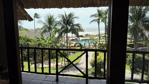 A view from a balcony with a thatched roof, overlooking a pool area, palm trees, and a beach. - Aroma Beach Resort & Spa (Phu Thuy)