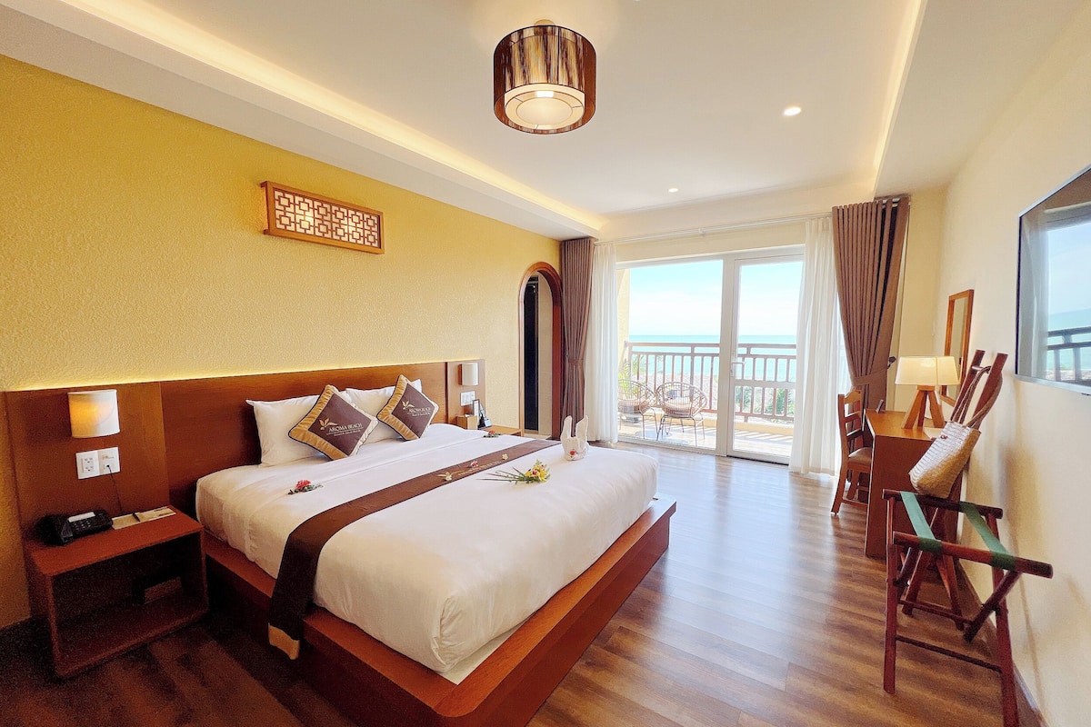 A hotel room with a large bed, a desk with chairs, a balcony with a view, and a ceiling light.