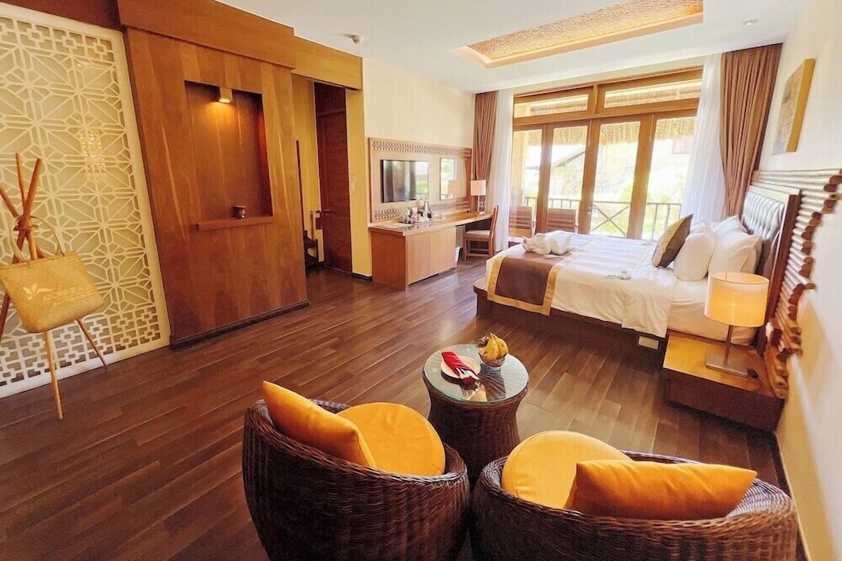 A hotel room with a bed, a desk, a TV, wicker chairs, and a decorative wall.