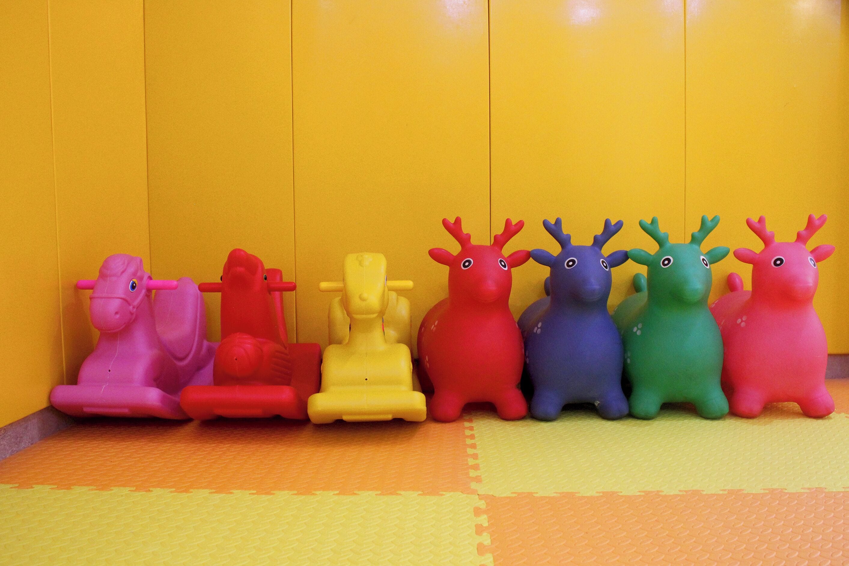 Une rangée d’animaux enjoués, de couleurs vives, est alignée sur un tapis devant un mur jaune.