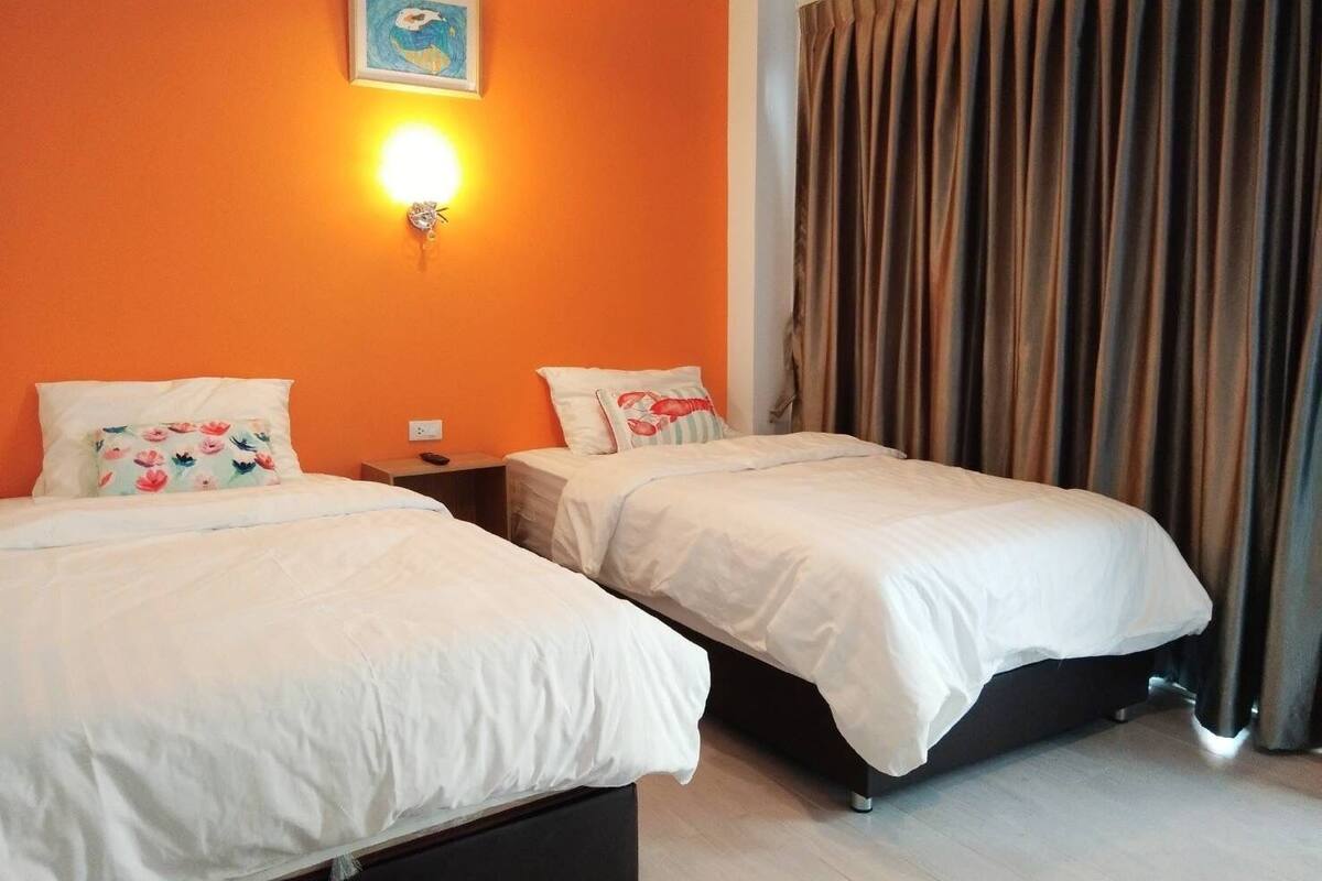A hotel room with two beds, an orange wall, a painting, a wall-mounted light, and a curtain.