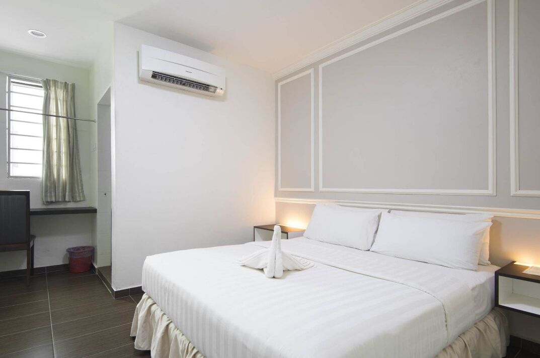 Habitación de hotel con cama, mesitas de noche, unidad de aire acondicionado, ventana con cortinas y un pequeño escritorio con silla.