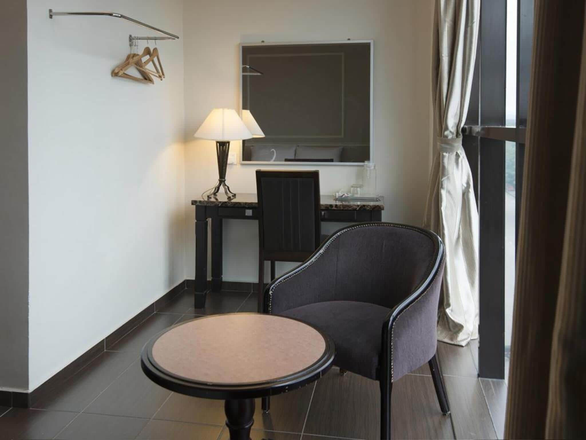 A hotel room with a round table, a chair, a desk with a lamp, a mirror, and a window with curtains.
