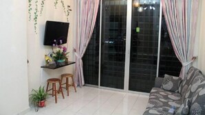 A living room with a sofa, a TV mounted on the wall, a small table with stools, and a sliding glass door with curtains. - Putra Place Home Inn (George Town)