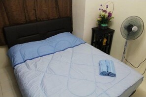 A bed with a blue quilt, a dark headboard, a small table with a fan and a vase of flowers, and a rolled towel on the bed. - Putra Place Home Inn (George Town)