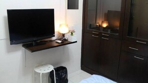 A hotel room with a flat-screen TV mounted on the wall, a bedside table with a lamp, a small stool, and a wardrobe with multiple doors. - Putra Place Home Inn (George Town)