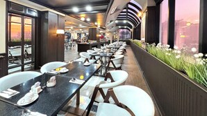 Un restaurant moderne avec de longues tables, des chaises blanches et une vue sur le paysage urbain grâce à de grandes fenêtres.