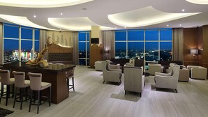 Un hall d’hôtel moderne doté d’un bar central, d’un espace salon et de grandes fenêtres offrant une vue sur la ville.