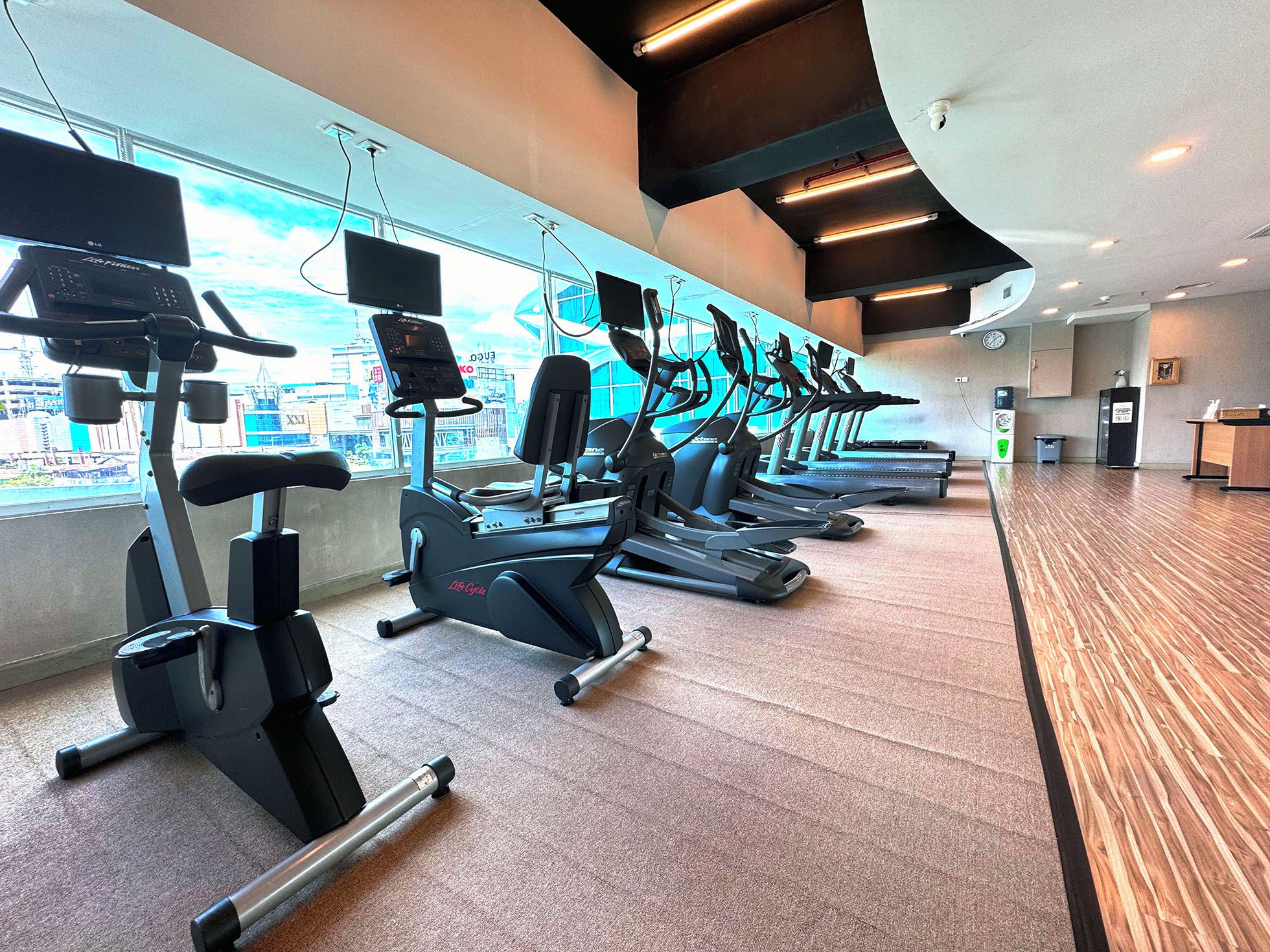 A gym with multiple treadmills, ellipticals, and exercise bikes, large windows with a city view, and a reception desk.
