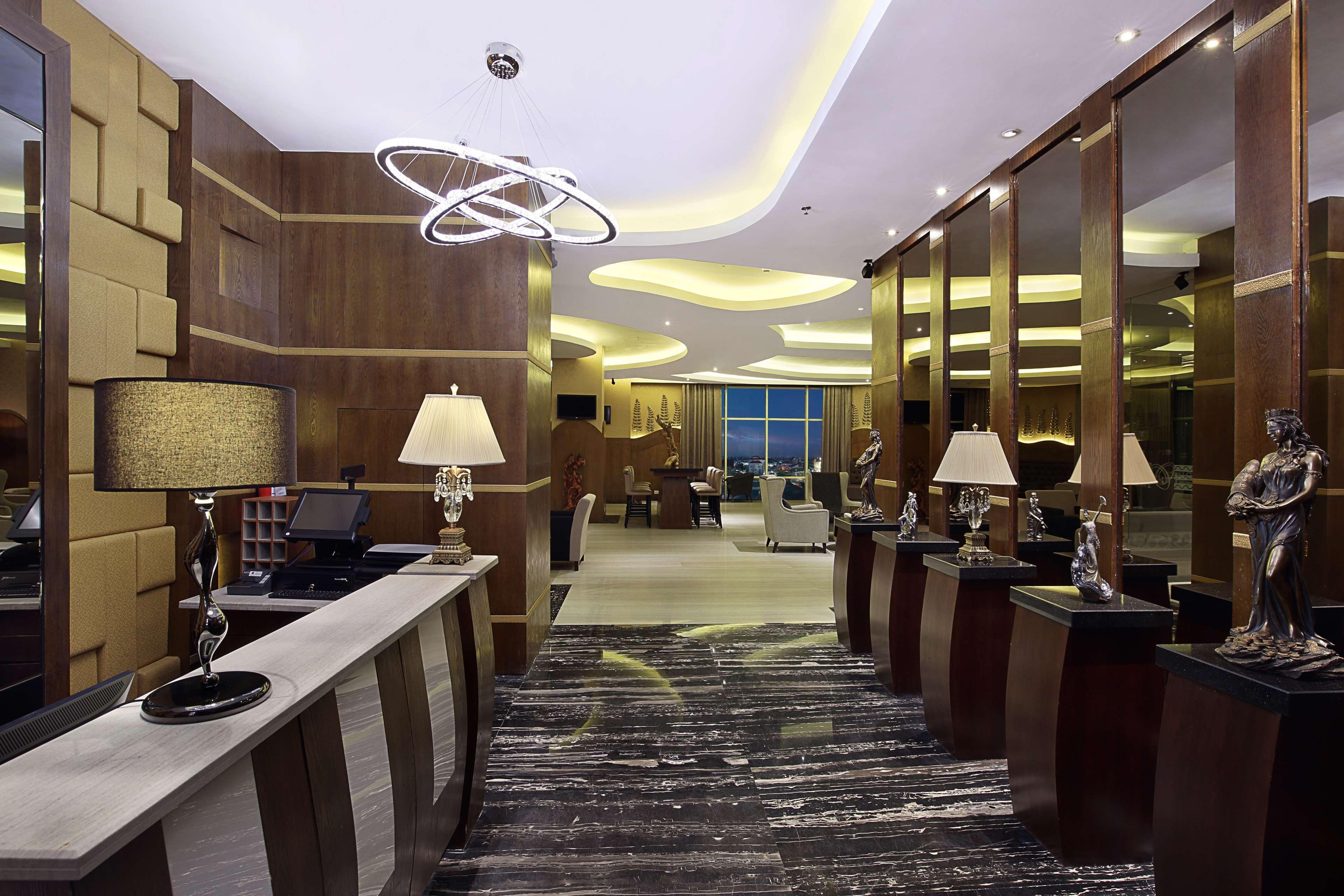 A hotel lobby with a reception desk, multiple lamps, a patterned carpet, and a large mirror.
