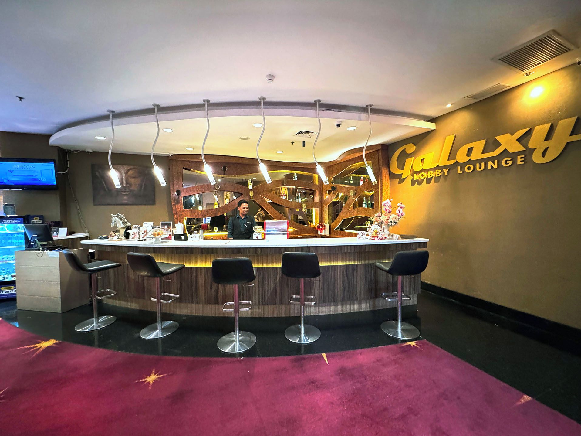 Un espace bar comprenant un comptoir, des tabourets de bar, un écran de télévision et un mur sur lequel est inscrit le texte « Galaxy Hotel Lounge ».