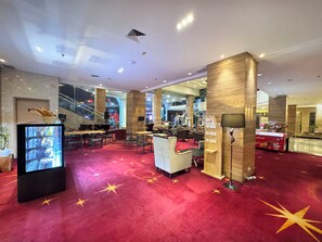 Vestíbulo de hotel con alfombra roja, zona de estar, un piano y una pantalla grande mostrando un paisaje urbano.