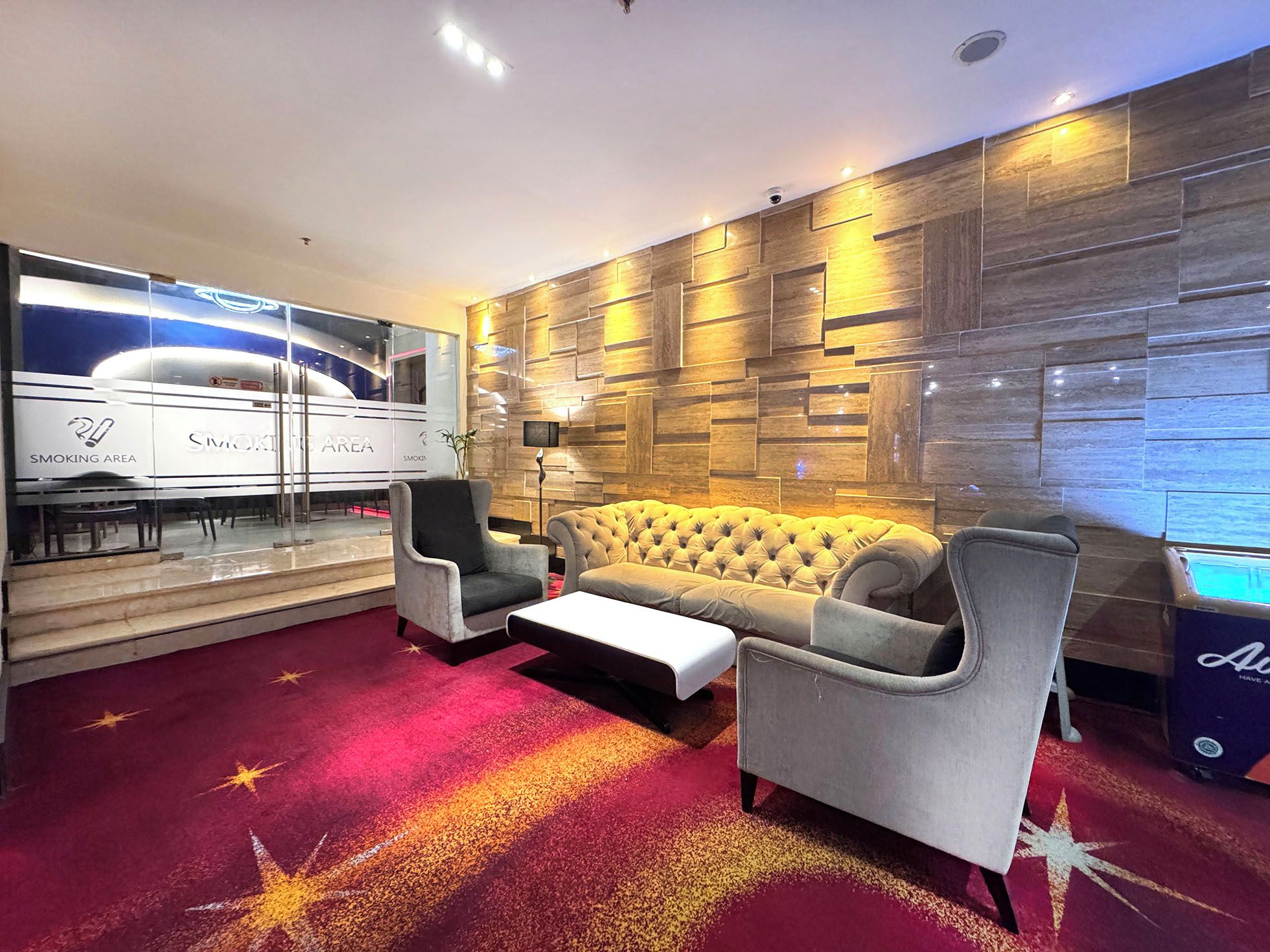 A modern lounge with a sofa, armchairs, a coffee table, a glass wall with a smoking area, and a wooden wall with recessed lighting.
