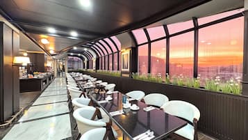 Area ristorazione di un hotel moderno con un lungo tavolo, sedie bianche e vista sulla città al tramonto.