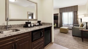 Habitación de hotel con cocina equipada, que incluye encimera de granito, fregadero, microondas y lavavajillas. Cuenta con un área de descanso con sofá y sillón, además de un ventanal con cortinas.