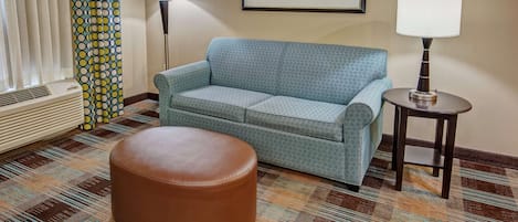 A room with a patterned carpet, a blue sofa, a brown ottoman, a standing lamp, and a radiator.
