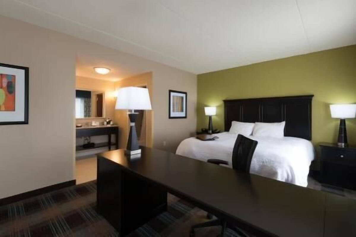 A hotel room with a bed, desk, chair, and a bathroom with a sink and mirror.
