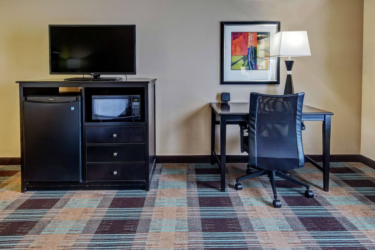 A hotel room with a flat-screen TV, a microwave, a refrigerator, a desk with a chair, a framed picture on the wall, and a patterned carpet.