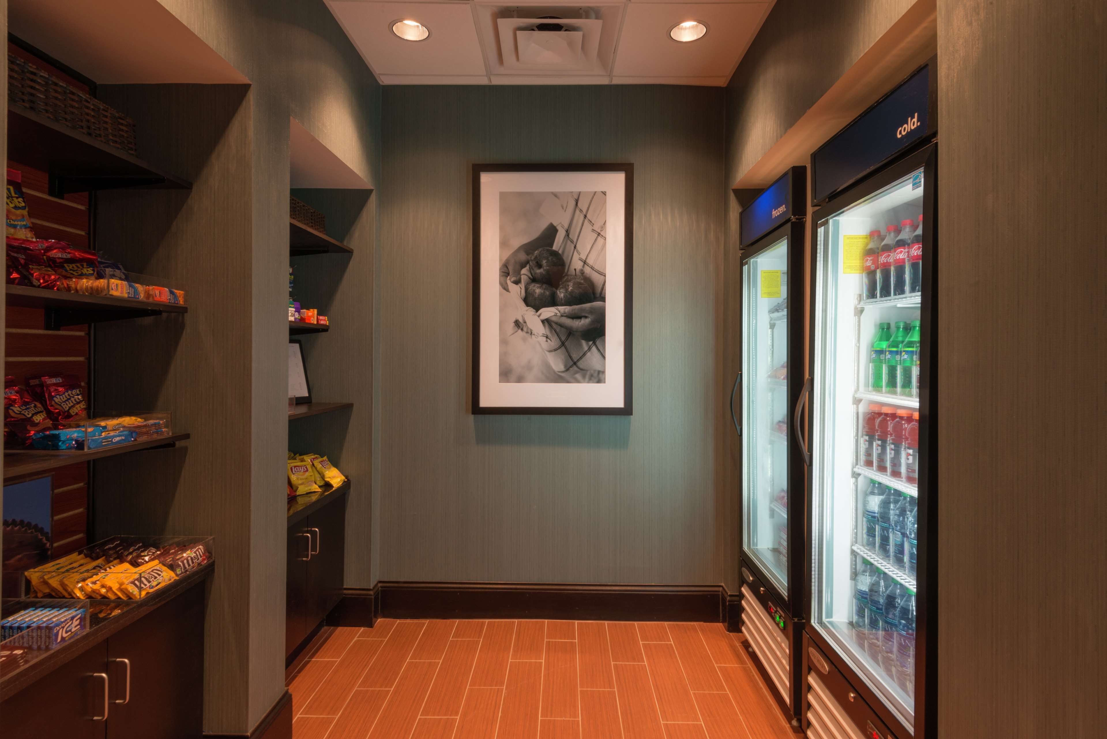 A narrow hallway with shelves stocked with snacks, a framed black and white photograph on the wall, and a refrigerator on the right.
