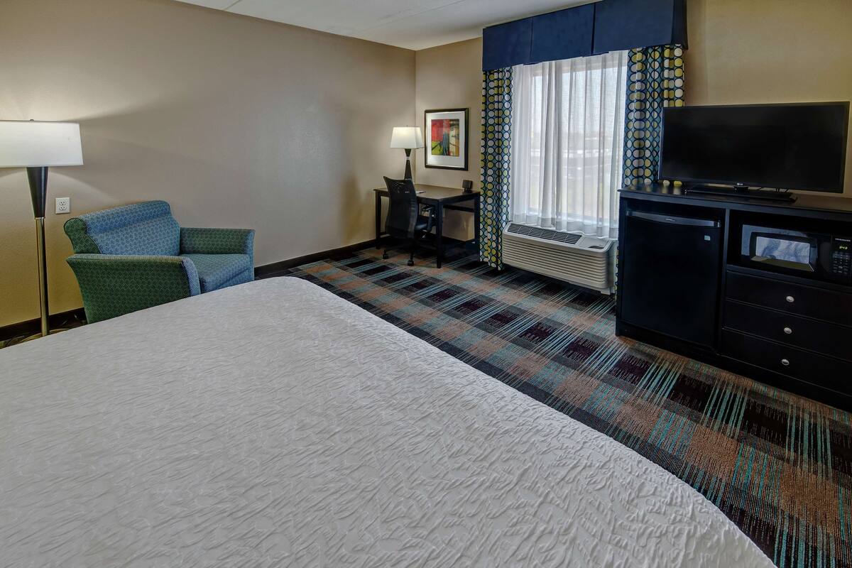 A hotel room with a large bed, a desk with a chair, a television, a microwave, and a window with curtains.