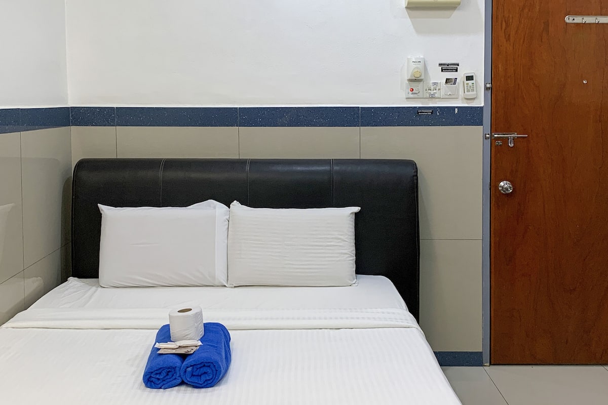 A hotel room with a bed, a wooden door, a wall-mounted thermostat, and a small table with toiletries.