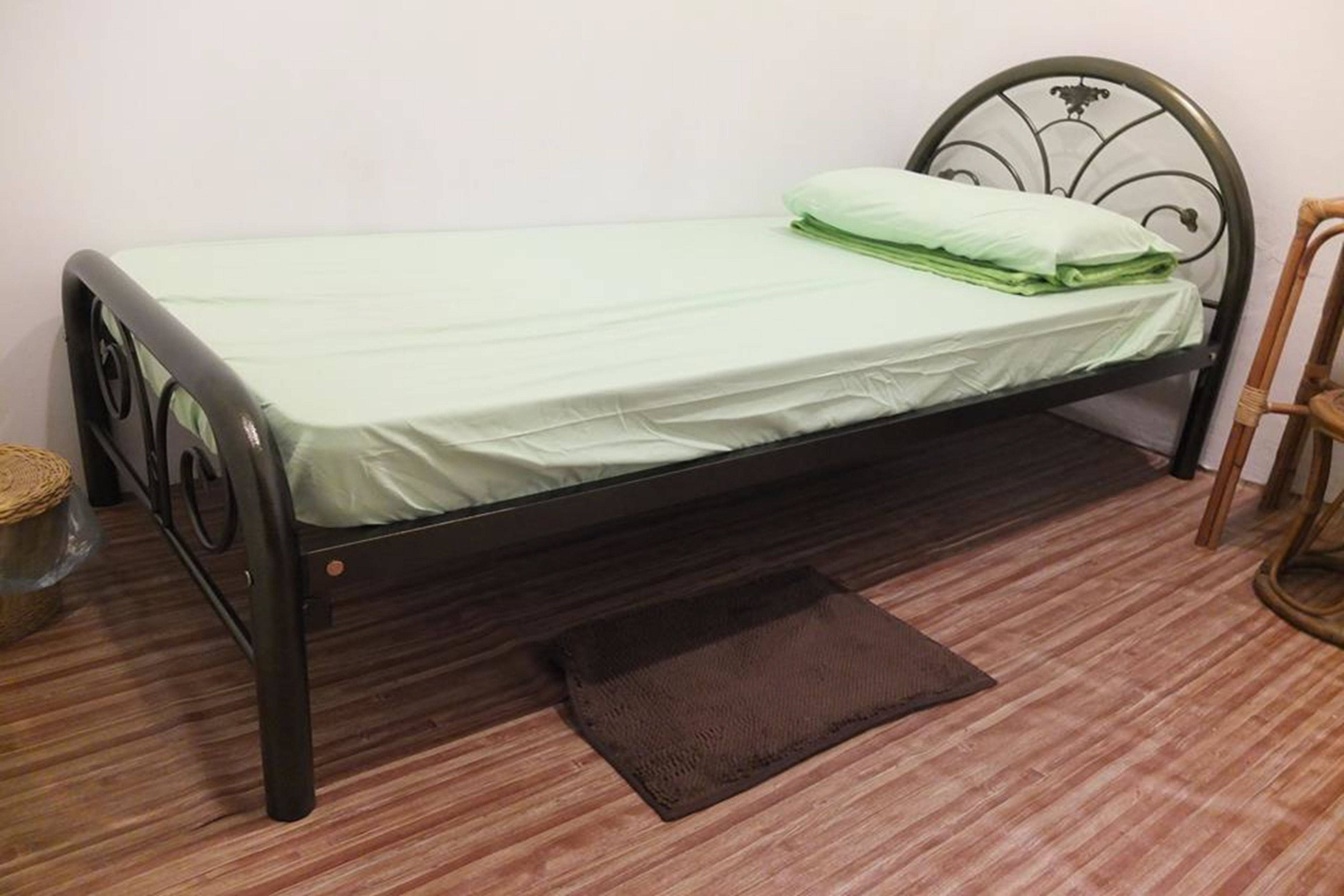 Una cama individual con estructura metálica, un colchón verde y una almohada a juego. Junto a la cama, hay un tapete marrón.