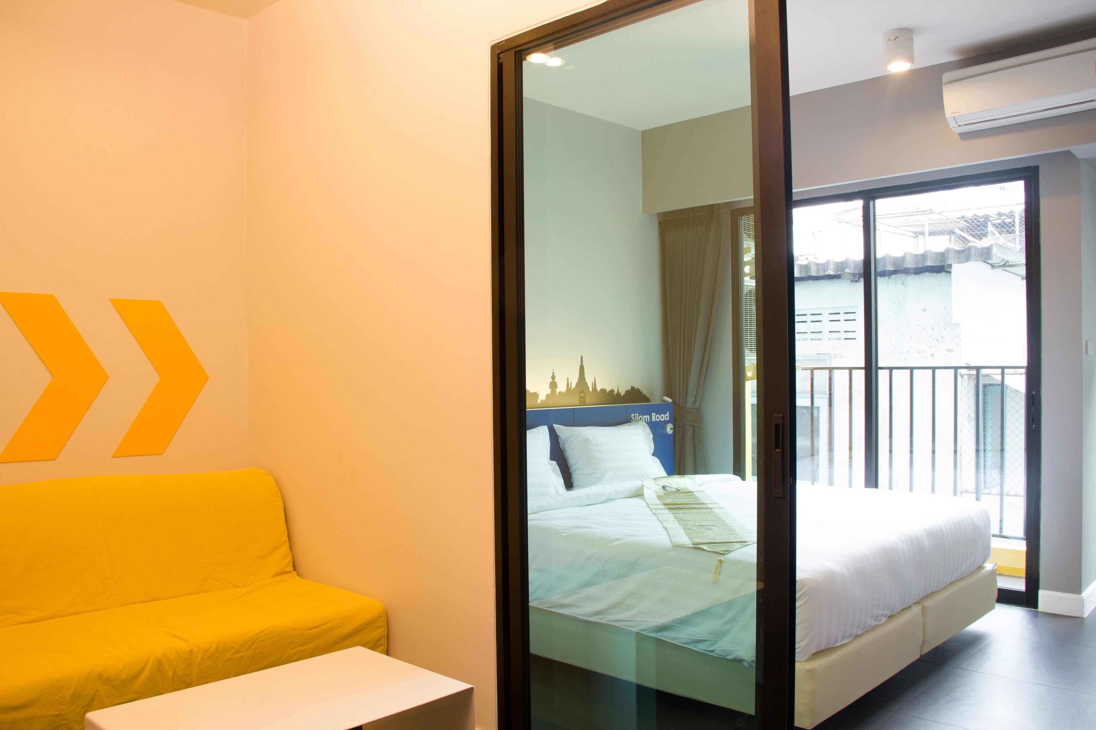 Una habitación de hotel con una cama, un sofá amarillo, una mesita y un ventanal que da a un balcón con vistas a la ciudad.