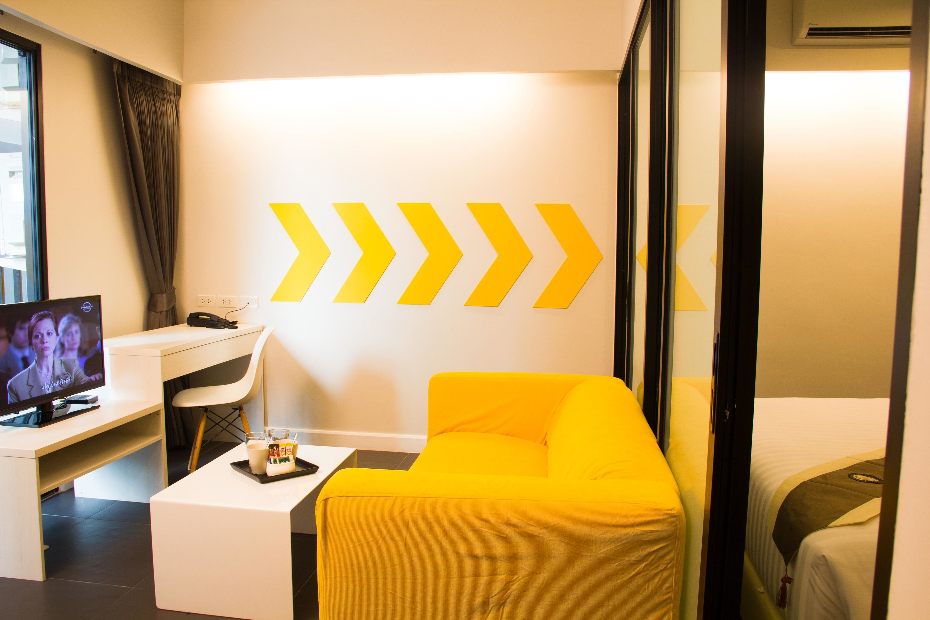 A hotel room with a yellow sofa, a desk with a computer, a television, and a bed.