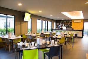 A modern restaurant with multiple tables and chairs, a bar area, and large windows providing natural light.