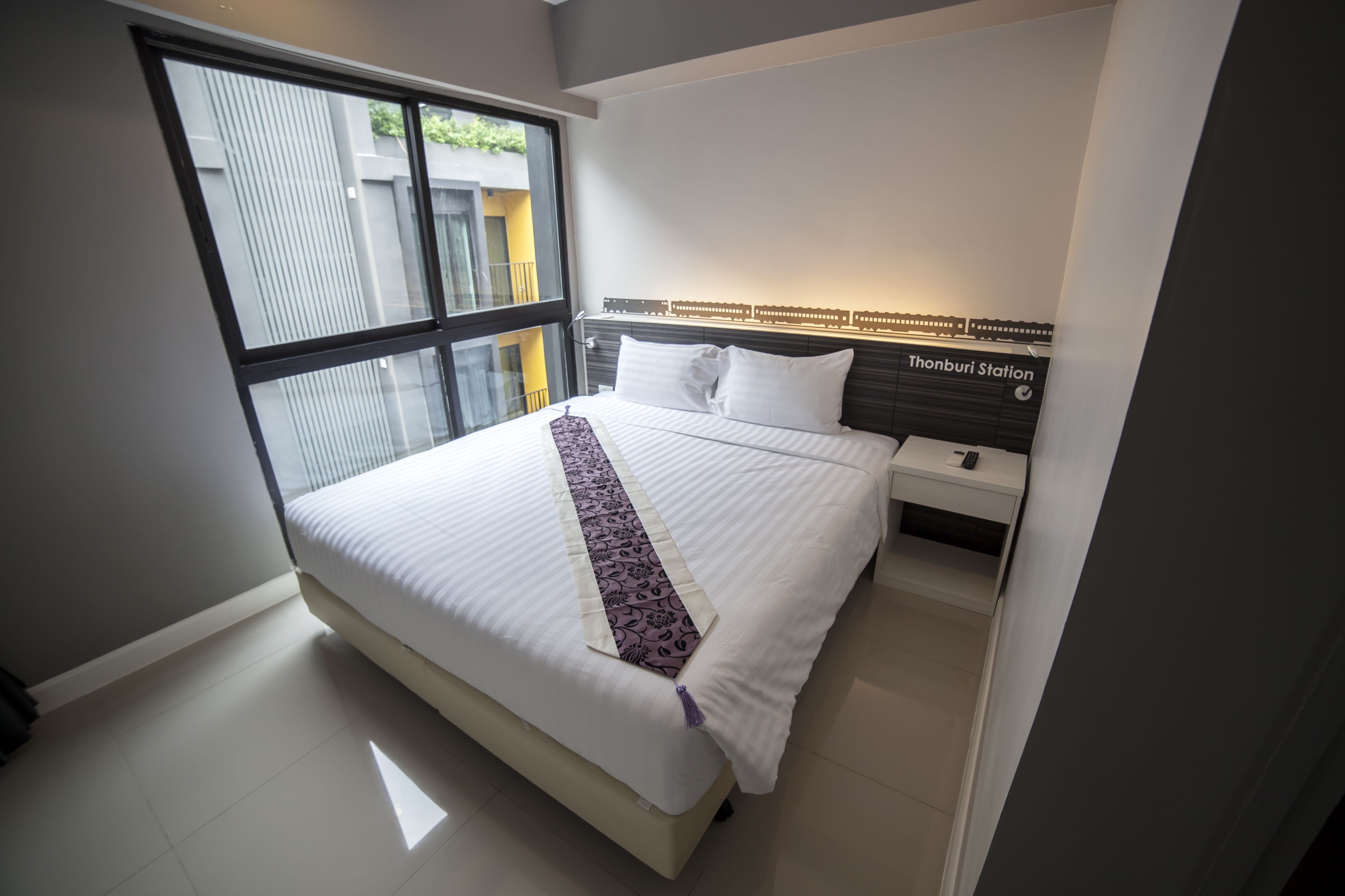 A modern hotel room with a large bed, a bedside table, a nightstand, and a view of the outside through a large window.