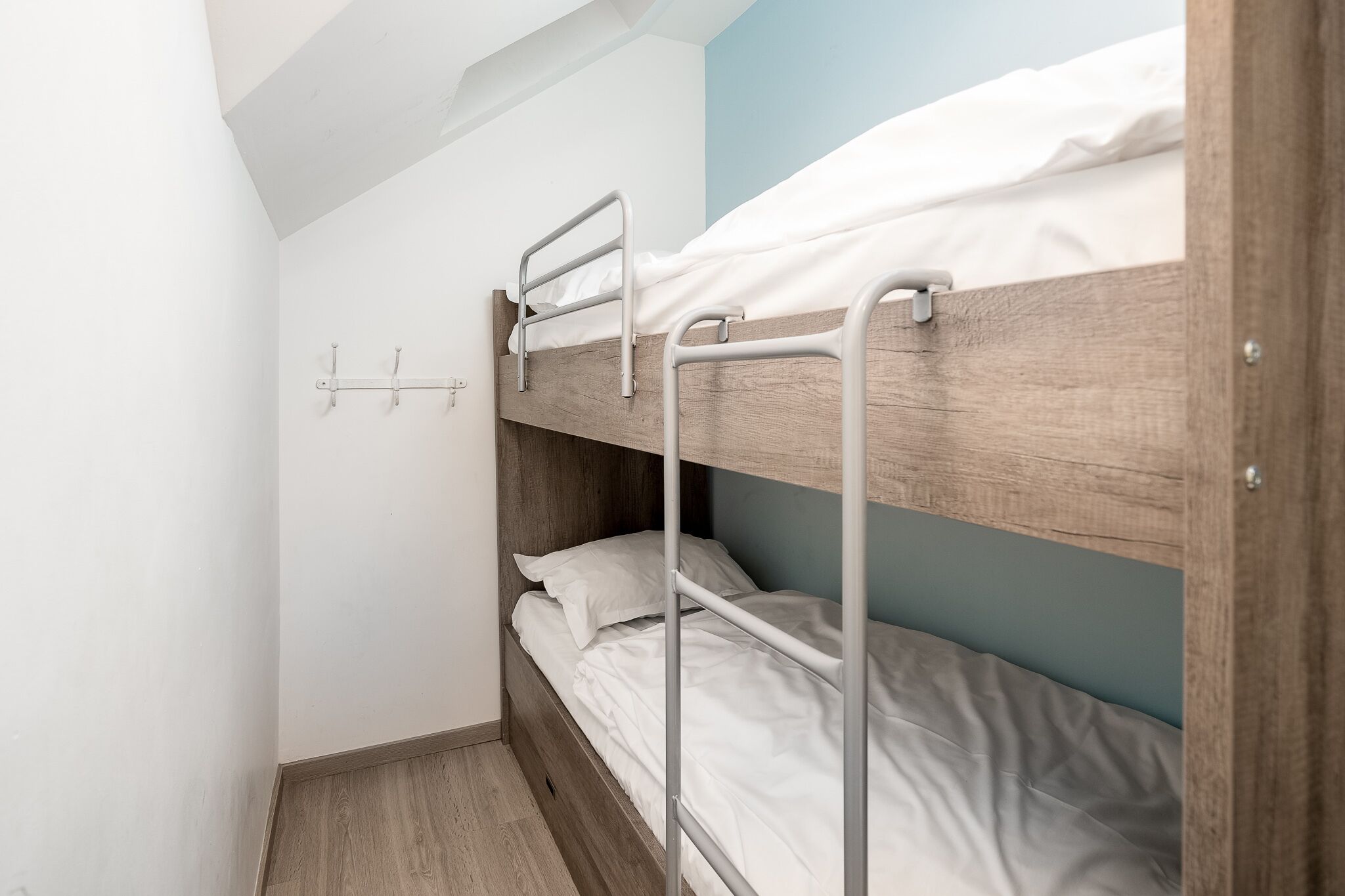 Ein Stockbett mit Metallgitter und weißer Bettwäsche in einem Zimmer mit hellblauen Wänden und Holzboden.