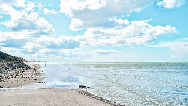 Una playa con costa rocosa, agua tranquila y cielo despejado con nubes.