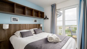 Habitación de hotel con cama, cabecera de madera, dos lámparas fijadas a la pared, ventana con cortinas y balcón con vistas.