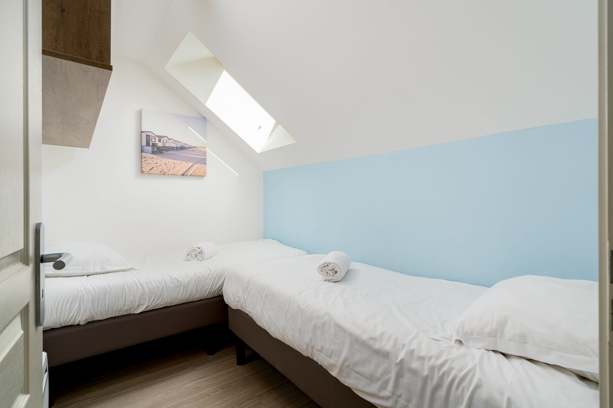 A bedroom with two beds, a skylight, and a painting on the wall.